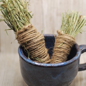 May include: Two bundles of dried grass, tied with twine, are in a blue ceramic mug. The mug has a dark blue glaze with a speckled pattern.