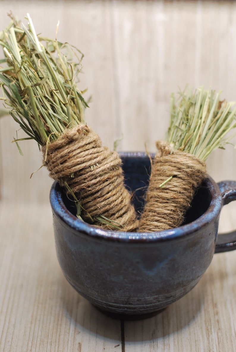 May include: Two bundles of dried grass, tied with twine, are in a blue ceramic mug. The mug has a dark blue glaze with a speckled pattern.