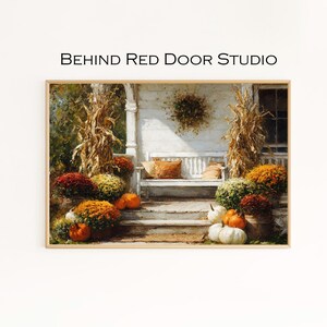 Könnte beinhalten: Ein gerahmter Druck einer Herbstszene mit einer weißen Veranda, einer Bank und Kürbissen. Das Bild zeigt bunte Chrysanthemen, Maisstängel und den Text "BEHIND Red Door STUDIO". Das Gesamtthema ist Herbst und Heimdekoration.