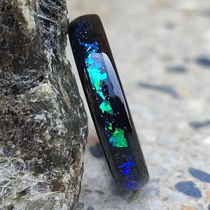 May include: A black ring with embedded iridescent green and blue glitter. The ring is sitting on a grey rock.