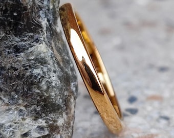 May include: A gold-colored ring leans against a gray rock. The ring has a smooth, polished surface and a simple, elegant design. The ring is likely made of gold or a gold-colored metal. The background is a light gray surface.