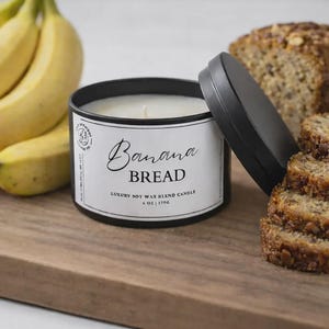 May include: A black metal container candle with a white label that reads "Banana Bread". The candle is a luxury soy wax blend, 6 oz / 170g. Sliced banana bread and bananas are also in the image, on a wooden surface.