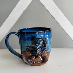 May include: A ceramic mug featuring a blue and brown glaze with a raised, three-dimensional black scorpion sculpture on the front. The mug has a curved handle and a textured base with white speckles, showcasing a blend of dark blue and earthy tones.
