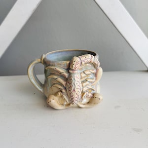 May include: A ceramic mug featuring a sculpted, three-dimensional scorpion relief on the exterior. The mug has a glossy, multi-toned glaze in shades of blue, tan, and cream, with a curved handle and a textured, detailed scorpion body design.