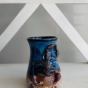 May include: A ceramic mug featuring a dark blue glazed upper section and a textured, crackled brown base. A sculpted, three-dimensional dark brown scorpion is attached to the front, with its tail curved upward and claws resting near the bottom.