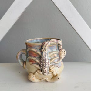 May include: A ceramic mug featuring a sculpted, three-dimensional scorpion relief on the exterior. The mug has a multi-colored, iridescent glaze with shades of blue, purple, and tan. It includes a curved handle and a detailed scorpion figure.