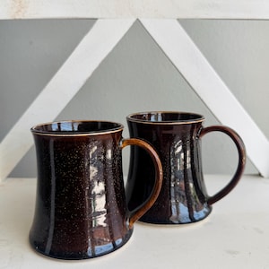 May include: Two dark brown ceramic mugs with a glossy finish and subtle light speckling. Each mug features a flared base, a curved handle, and a smooth rim. The mugs are positioned side by side, showcasing their deep brown color and reflective surface.