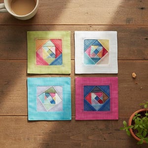 May include: Four square coasters with colorful geometric designs. Each coaster has a different colored border: green, white, blue, and pink. The coasters are arranged on a wooden surface, with a cup of coffee in the top left corner.