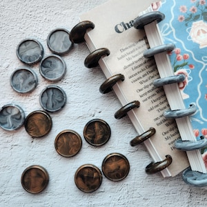 May include: An assortment of decorative wax seals in shades of gray and bronze, scattered around a notebook with a floral design. The notebook is held together with dark brown and gray disc-bound rings. The text "Cha" is visible on the page.