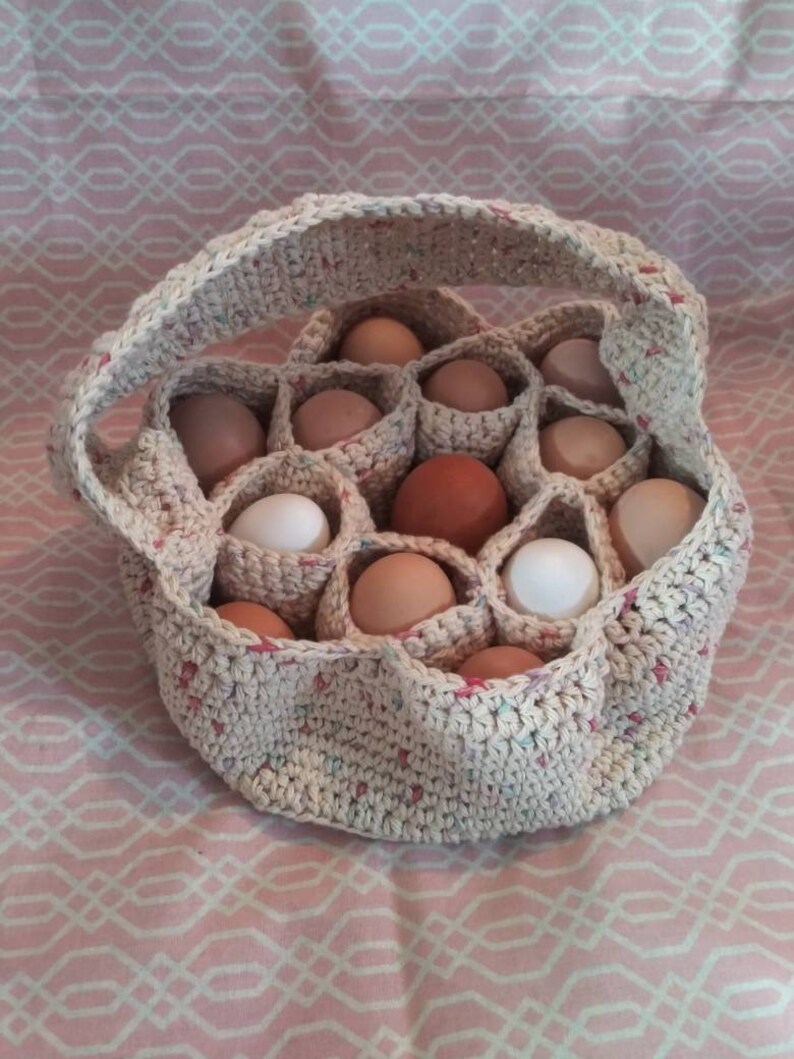 Puede incluir: Una cesta de huevos de ganchillo beige, llena de huevos de varios tonos marrones y blancos. La cesta tiene un asa y est&aacute; sobre una superficie rosa estampada. La cesta est&aacute; hecha de hilo con motas de color.
