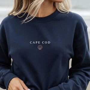 May include: Navy blue sweatshirt with the text "CAPE COD" in white, and a seashell graphic. The sweatshirt is a crew neck style. The model is wearing rings on their fingers.