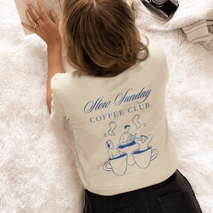 May include: A person wearing a beige t-shirt with a blue graphic of three people sitting in coffee cups. The text "Slow Sunday Coffee Club" is printed on the shirt.