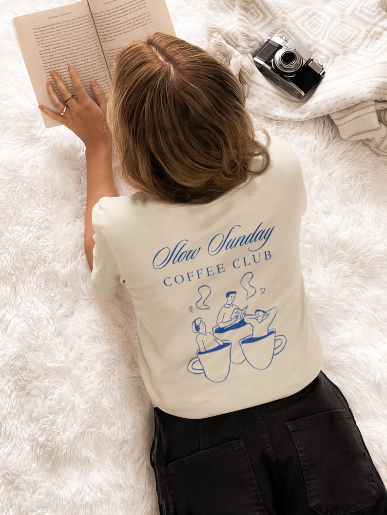 May include: A person wearing a beige t-shirt with a blue graphic of three people sitting in coffee cups. The text "Slow Sunday Coffee Club" is printed on the shirt.