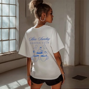 May include: White t-shirt with a blue graphic of three people sitting around a table with coffee cups. The text "Slow Sunday Coffee Club" is printed on the shirt.