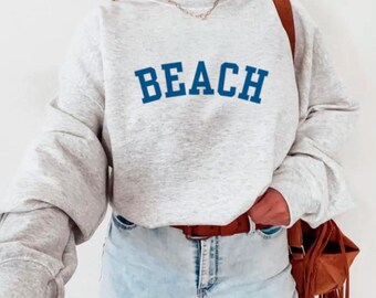 May include: A light gray sweatshirt with the word "BEACH" in blue block letters. The sweatshirt has long sleeves and is paired with light blue jeans and a brown belt. A brown shoulder bag with fringe is also visible.