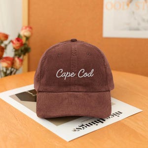 Puede incluir: Gorra de béisbol de pana marrón con el texto "Cape Cod" bordado en blanco. La gorra tiene una visera curva y una corona redondeada. El sombrero está sobre un trozo de papel.