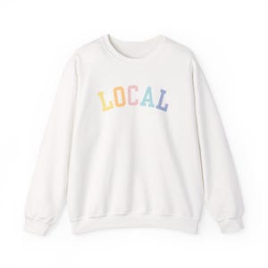 Puede incluir: Sudadera blanca de cuello redondo con la palabra "LOCAL" en colores pastel arcoíris en el pecho. La sudadera tiene mangas largas y un ajuste relajado. El diseño es simple y moderno, adecuado para ropa casual.