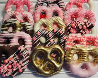 Könnte beinhalten: Eine Auswahl an Schokoladen-Brezeln mit verschiedenen Toppings, darunter rosa und weiße Streusel, Schokoladen-Drizzle und goldener Glitzer. Die Brezeln sind in einem durchsichtigen Plastikbehälter angeordnet.