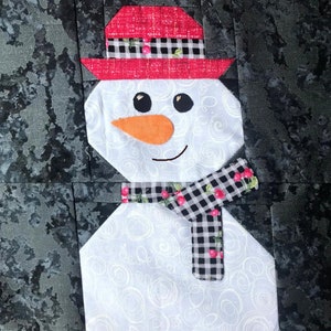 Puede incluir: Un muñeco de nieve blanco con un sombrero rojo y negro a cuadros, una bufanda roja y negra a cuadros con cerezas rojas y una nariz de zanahoria naranja.