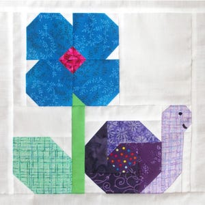 May include: A fabric block featuring a blue flower with a pink center and a green stem, next to a purple snail. The flower has a detailed leaf pattern. The snail has a patterned shell and a smiling face. The background is white.