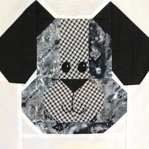 Puede incluir: Un bloque de patchwork con la cara de un perro. Las orejas del perro son negras, y la cara está hecha de una mezcla de tela a cuadros en blanco y negro y tela gris estampada. Los ojos son pequeños círculos negros, y la nariz es un triángulo negro.