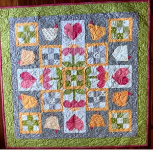 May include: A patchwork quilt with a gray background and a repeating pattern of hearts, flowers, and chickens. The quilt is made up of squares and rectangles of different colors and patterns, including green, yellow, pink, and white.