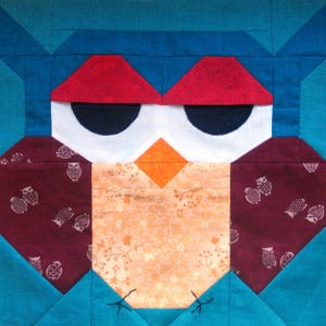 May include: A patchwork owl quilt block with a teal blue background, red eyebrows, white eyes with black pupils, an orange beak, and a peach-colored body with a floral pattern. The block is surrounded by smaller teal blue squares.