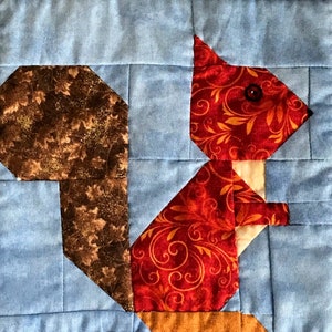 May include: A red and brown patchwork quilt block featuring a squirrel design. The squirrel is made of red fabric with a gold floral pattern. The tail is made of brown fabric with a speckled pattern.