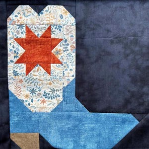 Puede incluir: Un bloque de patchwork con forma de bota de vaquero. La bota presenta una estrella color óxido sobre un fondo floral, con una caña azul y un talón marrón. El fondo es de color azul marino oscuro.