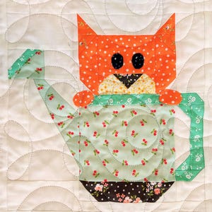 May include: A patchwork quilt block depicting an orange cat with black eyes and a floral face, sitting on a green watering can. The watering can has a cherry print and floral base, set against a white background with a swirling pattern. The quilt is machine stitched.