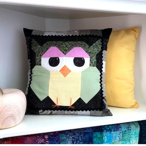 Könnte beinhalten: Ein grünes, gelbes, rosa und schwarzes Kissen in Form einer Eule mit einem schwarzen und weißen gewellten Rand. Das Kissen liegt auf einem weißen Regal mit einem gelben Kissen rechts.