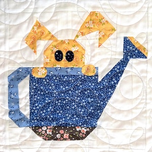 Op de afbeelding: Een patchwork vierkant met een gieter met een konijnengezicht. De gieter is blauw met een bloemenpatroon en het gezicht van het konijn is geel met zwarte ogen. De achtergrond is wit met een gewatteerd patroon.