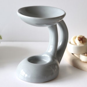 May include: Grey ceramic essential oil diffuser with a curved design. The diffuser has a small bowl on top for essential oils and a lower bowl for a tea light candle.