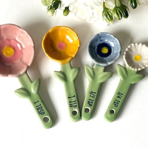Puede incluir: Juego de cuatro cucharas medidoras de cerámica con forma de flores en rosa, amarillo, azul y blanco. Cada cuchara tiene un tallo verde con diseño de hoja y una etiqueta de medida: 1 cucharada, 1 cucharadita, 1/2 cucharadita y 1/4 cucharadita.