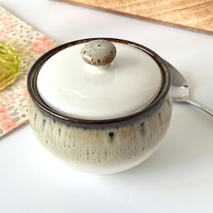 May include: A white ceramic sugar bowl with a lid and a brown rim. The bowl has a speckled glaze in shades of brown, beige, and white.