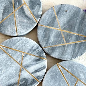 May include: Set of four round grey marble coasters with gold geometric lines. The coasters are approximately 10 cm in diameter.