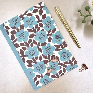 Könnte beinhalten: Ein Notizbuch mit blauem und braunem Blumenmuster, ein goldener Stift und zwei roségoldene Büroklammern. Das Notizbuch hat einen hellblauen Rand und ein sich wiederholendes Blumenmuster.
