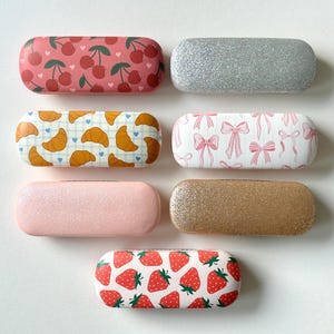 May include: Seven eyeglass cases with different patterns. The cases feature designs of cherries, croissants, bows, strawberries, and glitter. The cases are in pink, gold, and silver, with a white background.
