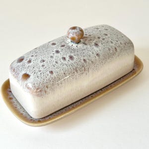 May include: A rectangular ceramic butter dish with a lid and a matching base. The dish is mainly white with a speckled, brown and grey glaze on the lid and sides. The base is a light brown colour. The lid has a small, round knob.