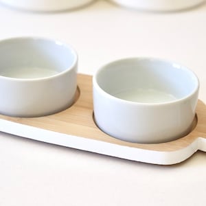 May include: A white ceramic serving tray with two small bowls, set on a wooden base with a white painted edge. The tray has a handle on the side.