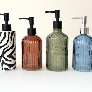 Könnte beinhalten: Vier dekorative Seifenspender mit schwarzen Pumpen. Der erste Spender hat einen schwarz-weißen Zebradruck. Der zweite ist eine gerippte, bernsteinfarbene Glasflasche. Der dritte ist eine grüne, gerippte Glasflasche mit dem geprägten Wort "SOAP". Der vierte ist eine blaue, gerippte Glasflasche.