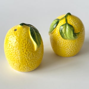 May include: A set of two ceramic lemon salt and pepper shakers. The shakers are yellow with green leaves and are shaped like lemons.