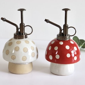 May include: Two mushroom-shaped plant misters. One is white with tan polka dots, the other is red with white polka dots. Both have a bronze-colored sprayer and handle. The base is a light tan color.