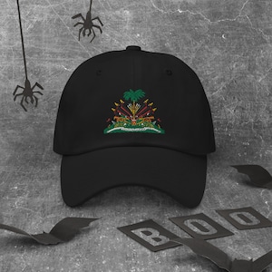 May include: Black baseball cap with an embroidered patch featuring the coat of arms of Antigua and Barbuda. The patch is green, yellow, red, and white. The cap is on a gray background with black bats and spiders.