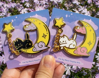 May include: Two enamel pins featuring a cat resting on a yellow crescent moon with a star and chain. One pin has a black cat, the other a white cat. The pins are on a blue card with the text "Meow Royale Art".