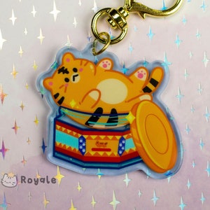 Op de afbeelding: Een gouden sleutelhanger met een bedel met een cartoon oranje kat die op een kleurrijke trommel ligt. De bedel is bevestigd aan een gouden ketting met een hartvormige sluiting.