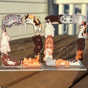 Op de afbeelding: Twee transparante acryl display stands versierd met cartoon katten. Elke standaard heeft een rechthoekig frame met kattenillustraties in verschillende kleuren en poses. De stands zijn ontworpen om kleine items vast te houden.