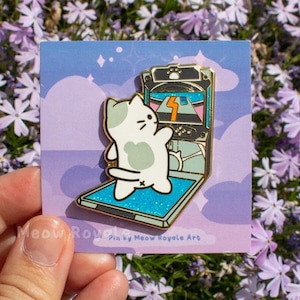 May include: Enamel pin featuring a white and gray cat playing an arcade game. The pin has a blue background with a cartoon cat and a retro arcade machine. The pin is on a square card with the text "Meow Rogue Art".