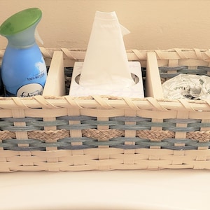 May include: A white wicker basket with blue and tan accents holds two spray bottles, a box of tissues, and a roll of toilet paper.