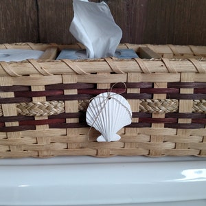 May include: A woven wicker basket with a brown and tan pattern. The basket has three compartments, one for a roll of toilet paper, one for a box of tissues, and one for other items. A white seashell is attached to the front of the basket.